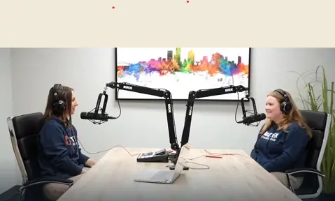 Two girls sitting across from each other taking into microphones in a podcast studio