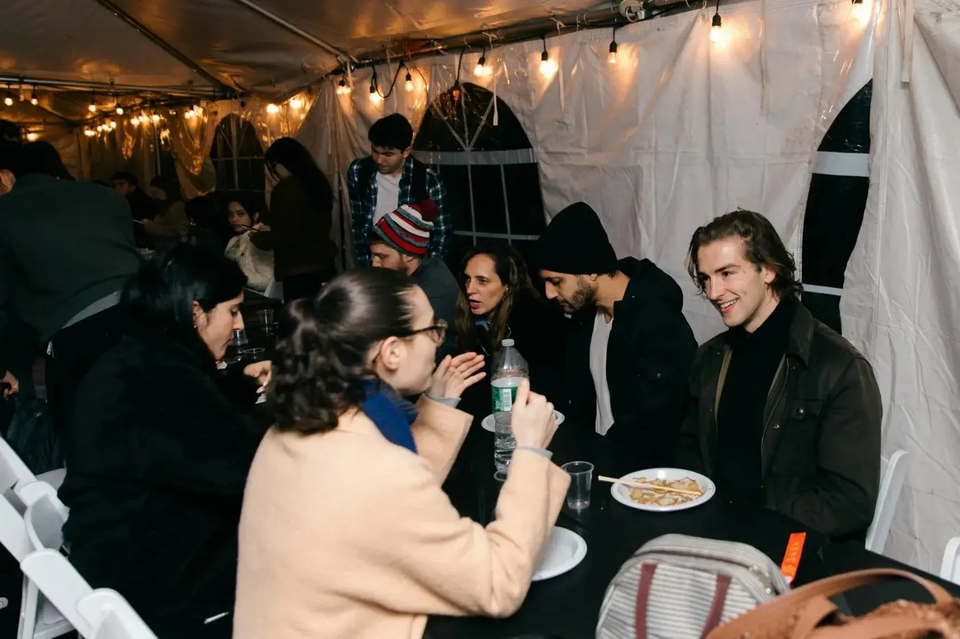 People sitting and talking at a gathering inside a tent with string lights, eating food and drinking from bottles and glasses.