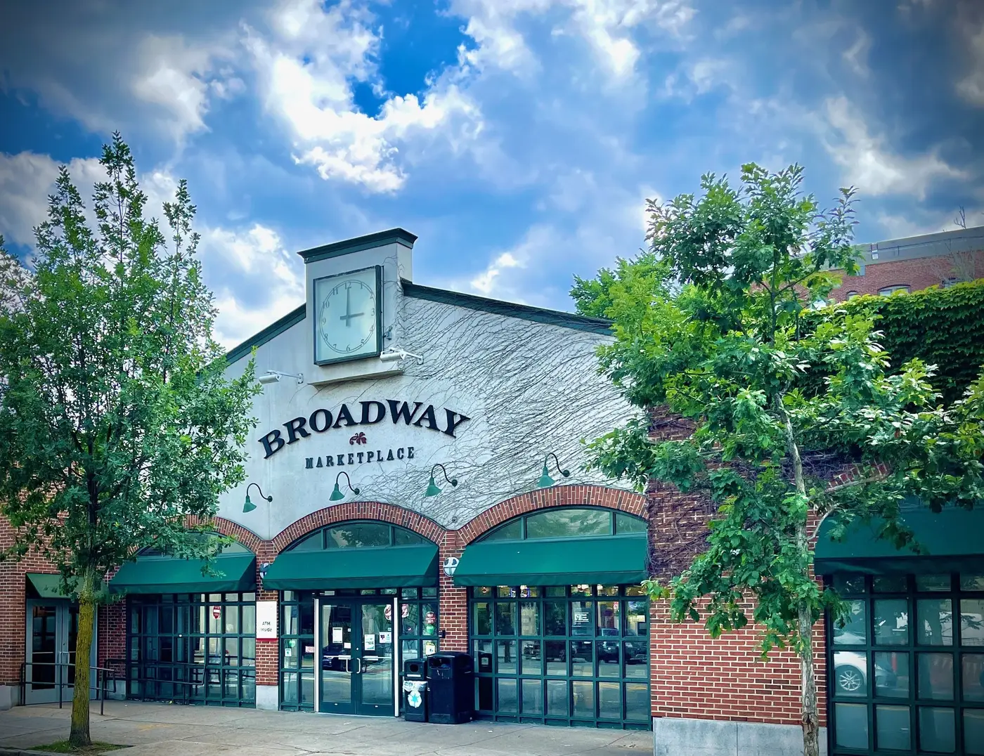 Exterior view of Broadway Marketplace with brick facade, green awnings, trees in front, and a blue sky with clouds.