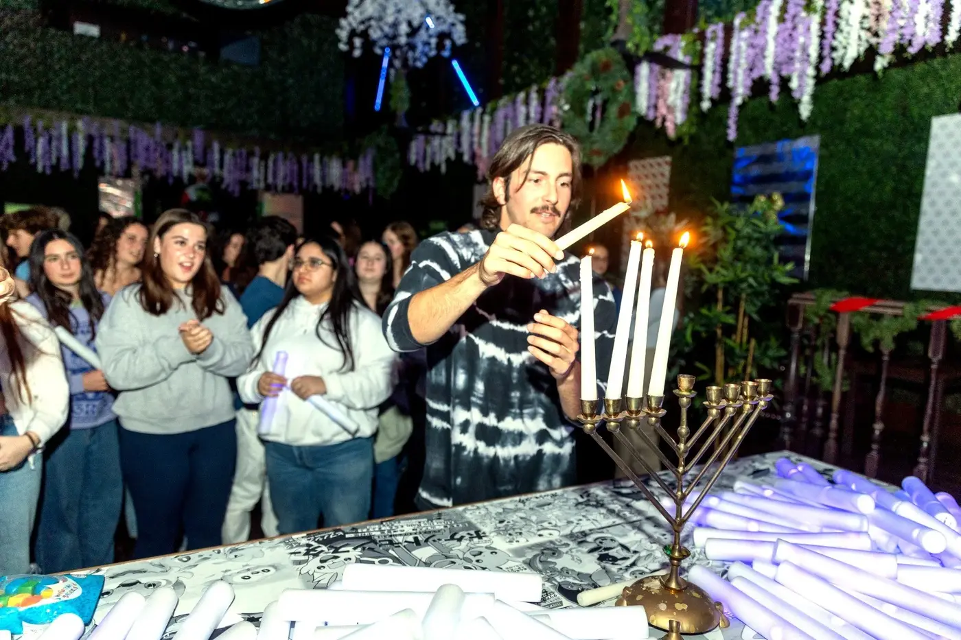 A man is lighting candles on a menorah during Hanukkah celebration, with a group of people watching.