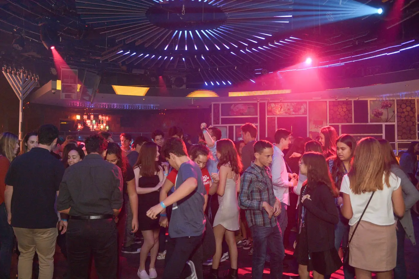 Group of young people dancing and socializing in a nightclub with colorful lights and a DJ booth in the background.