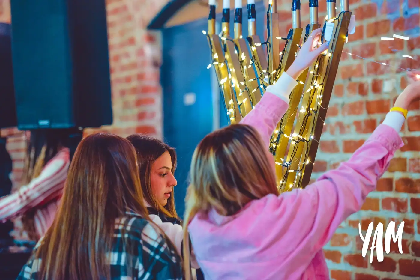 Three young women decorating a wooden stand with string lights in an indoor setting with brick walls.