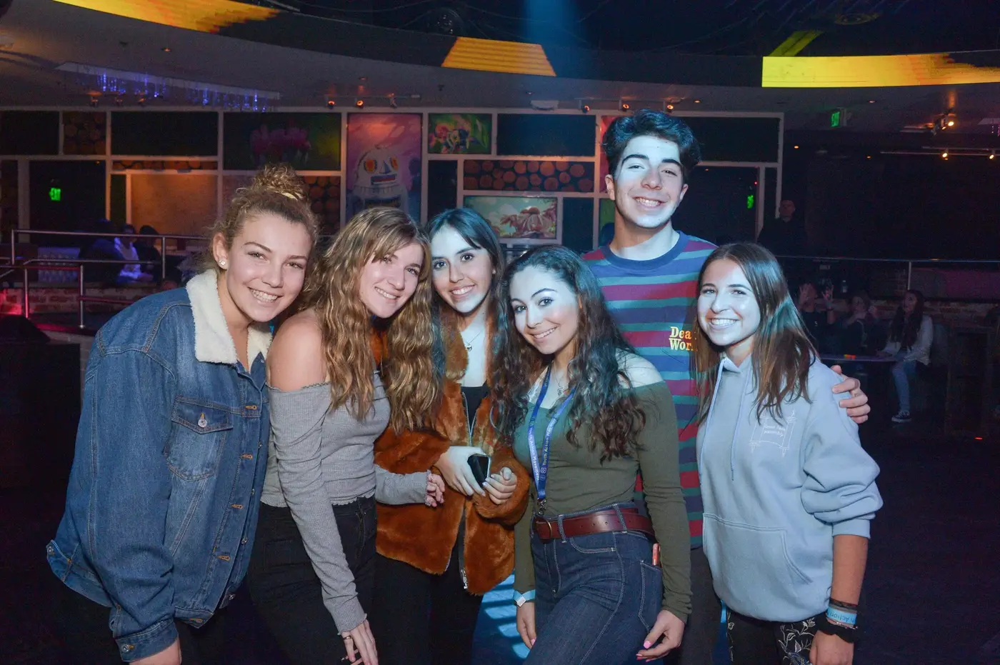 Group of six young people smiling and posing together in a colorful indoor entertainment venue with arcades and artwork in the background.