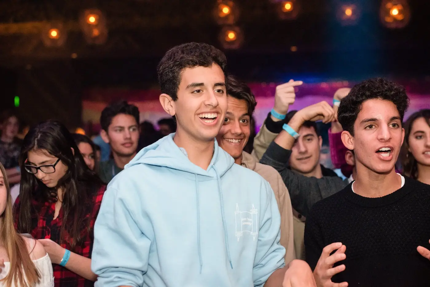 Group of young people enjoying a crowded event or concert, some smiling and talking, with colorful lighting in the background.