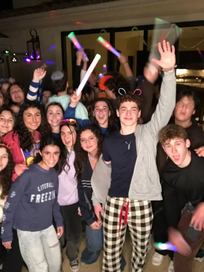 A group of teenagers at a party, smiling and cheering, surrounded by glow sticks and party decorations.
