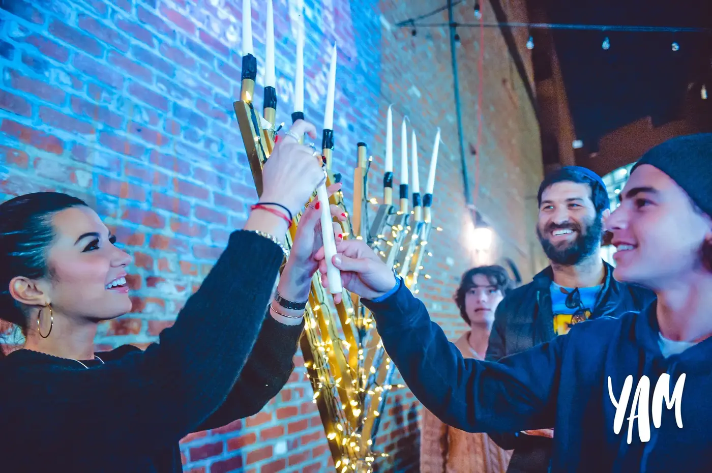 Group of four people celebrating, lighting candles on a menorah during Hanukkah, with a brick wall and festive lights in the background.