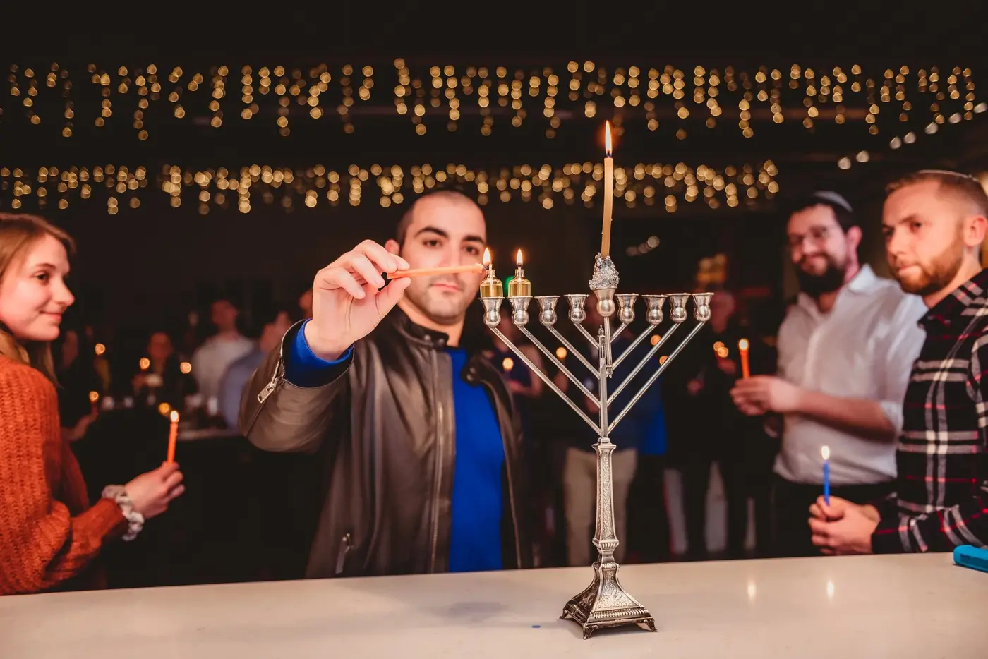 People lighting candles on a large menorah during a Hanukkah celebration in a decorated indoor setting.
