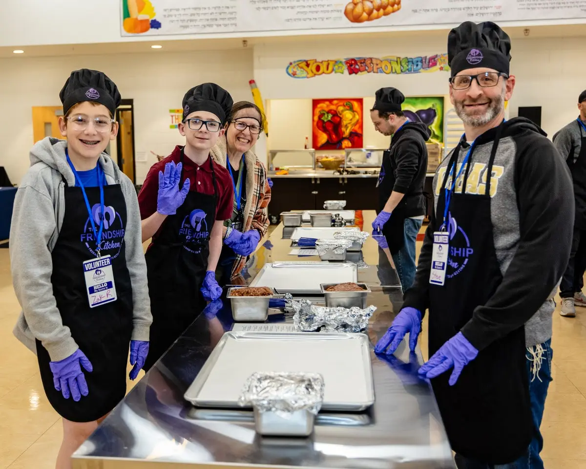 Four smiling people wearing black chef hats, aprons, and purple gloves stand behind a metal counter with food.