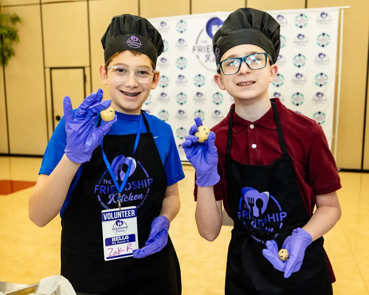 Two boys in chef hats, aprons, and purple gloves hold up small cookie dough balls, smiling at the camera.