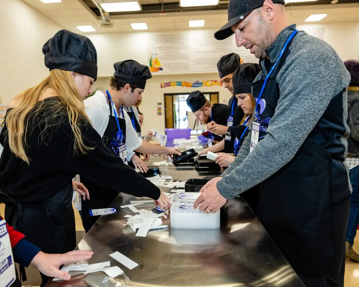 Several people in black chef hats and aprons work at a long metal table, packaging white strips.