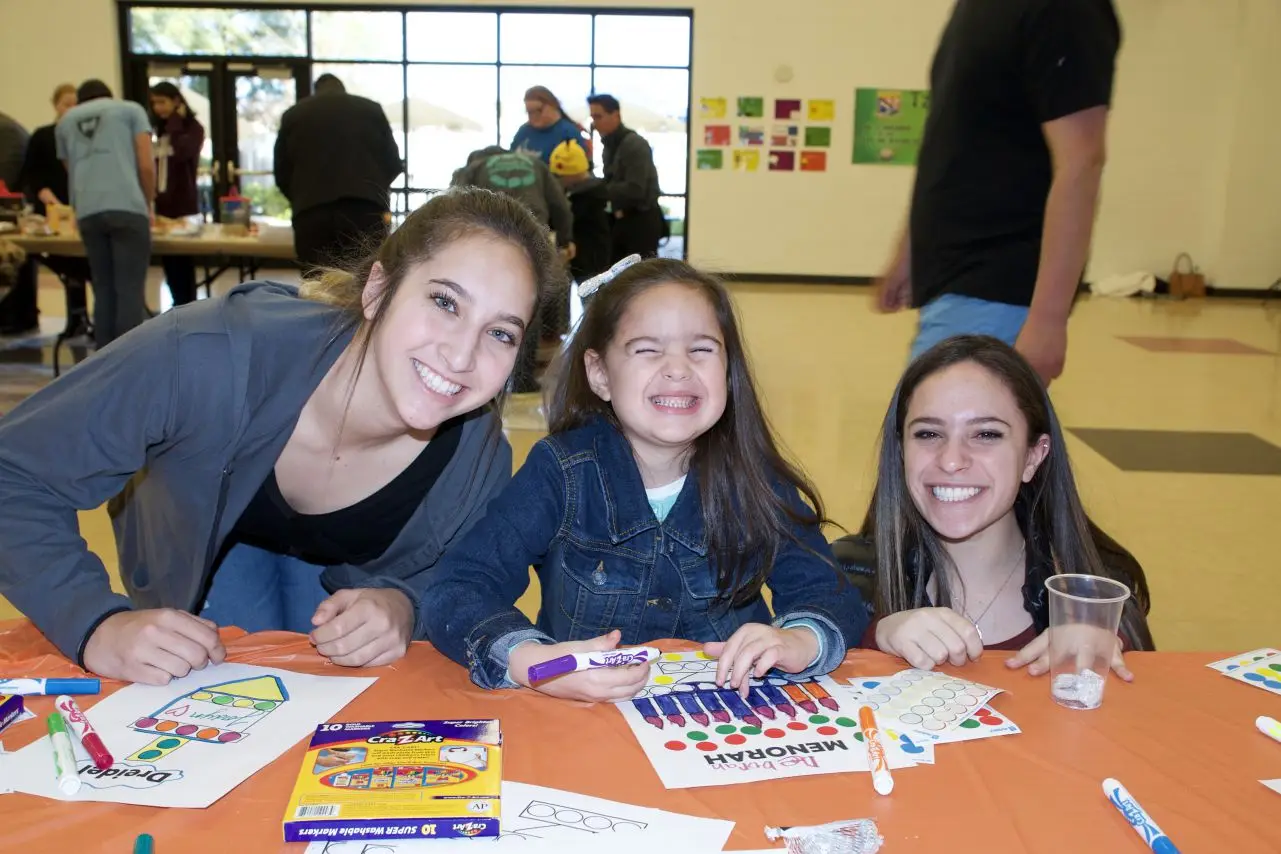 Three smiling girls, two teens and one child, sit at a table with markers and "MENORAH" coloring sheets.