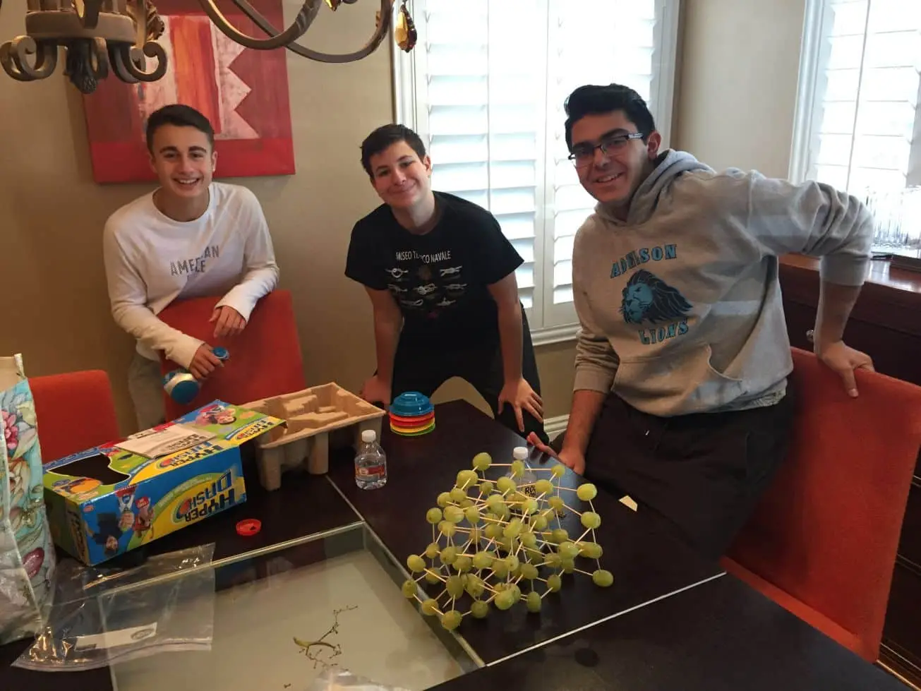 Three smiling young men stand around a table with a "Hyper Dash" box and a grape and toothpick structure.