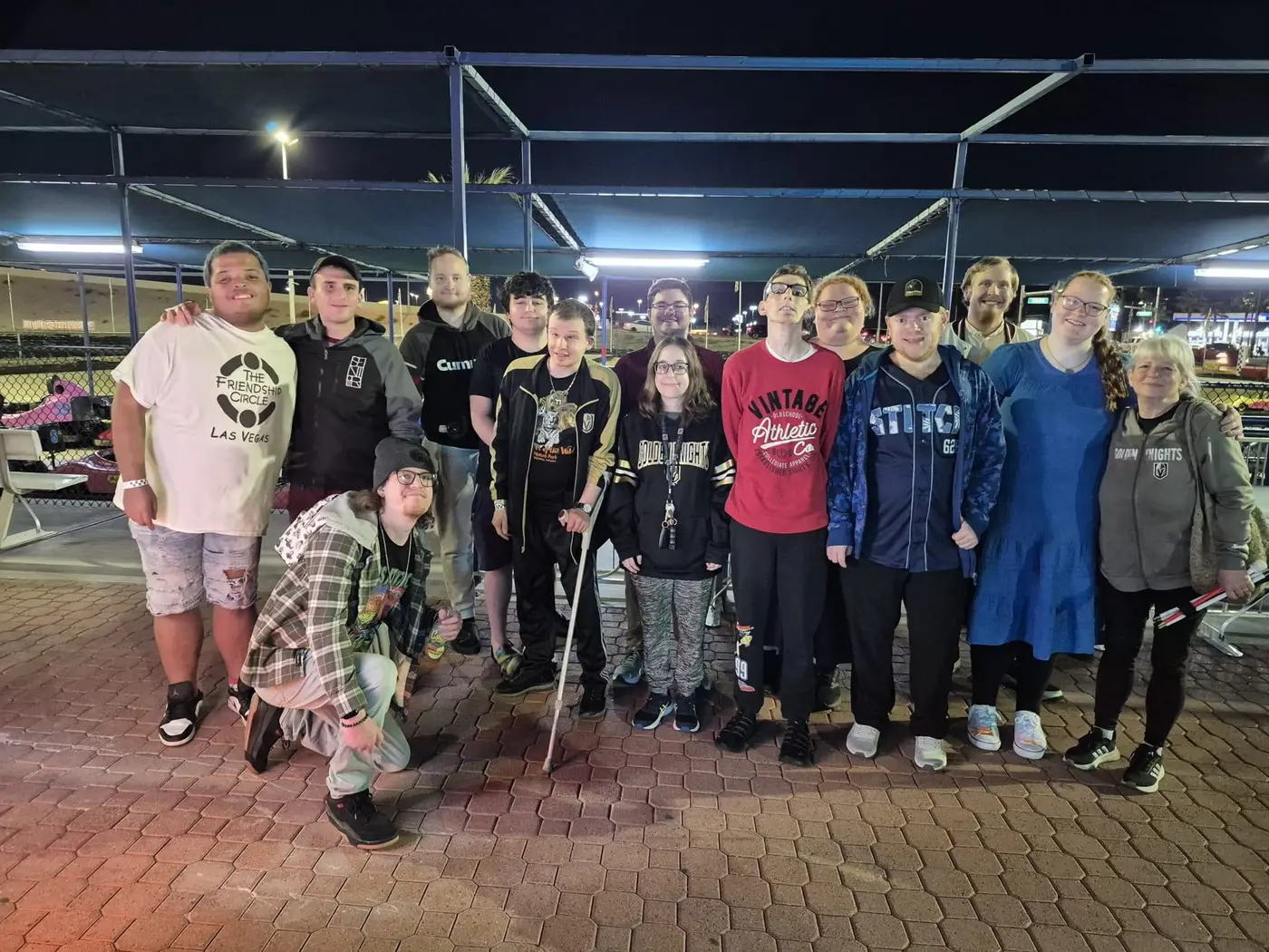 A diverse group of fifteen smiling people pose for a photo at night on a paved surface.