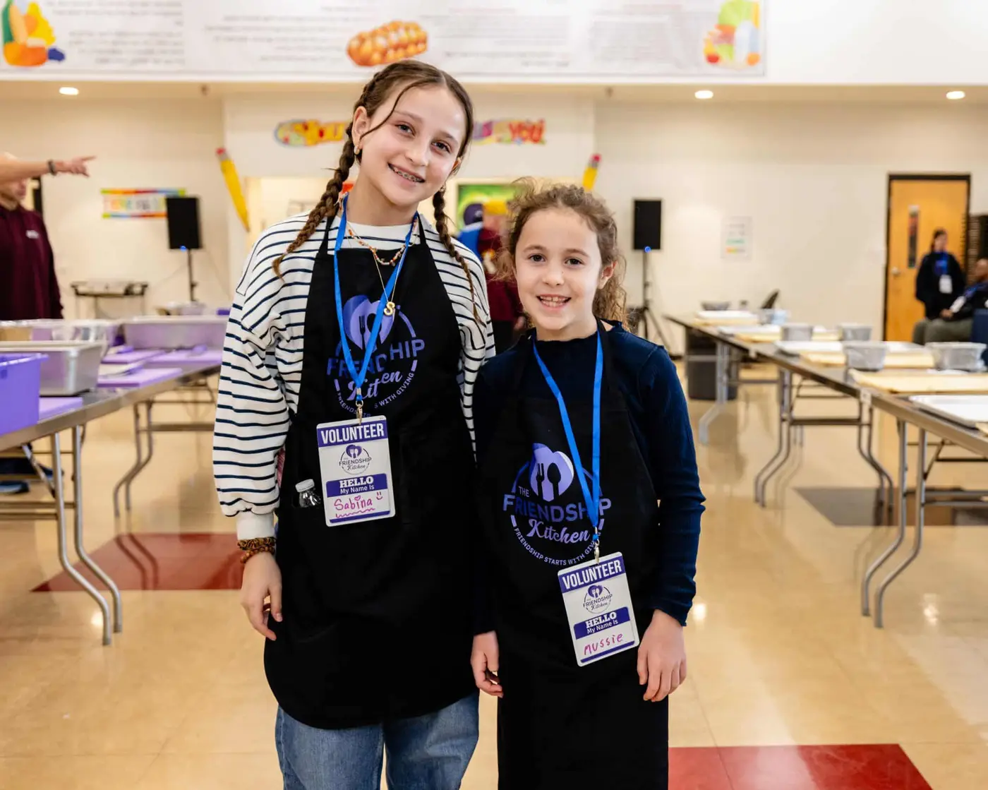 Two smiling girls in black aprons with "The Friendship Kitchen" logo and "Volunteer" name tags.