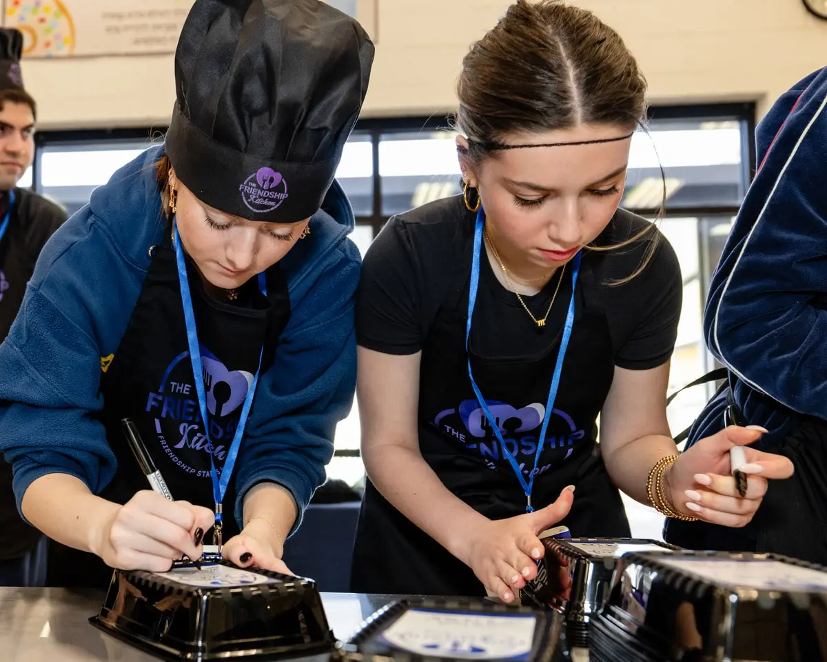 Two young women in aprons and lanyards, one in a chef's hat, write on black containers.