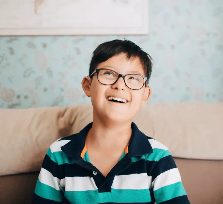 A young boy with dark hair and glasses wears a striped polo shirt, laughing and looking up.