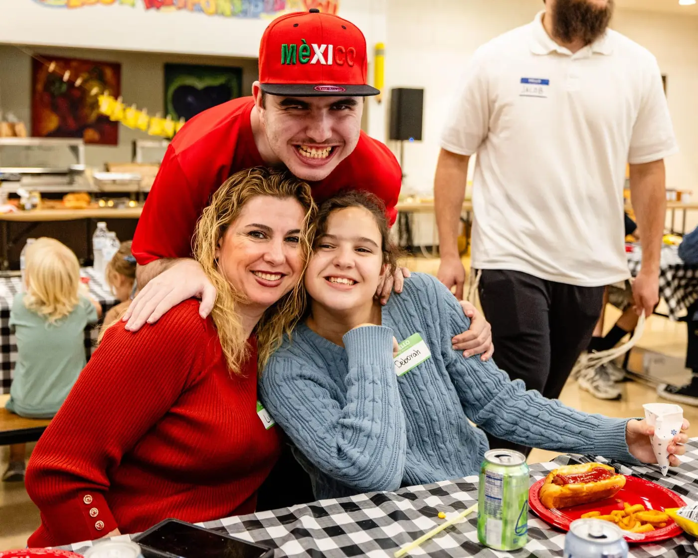 A smiling man in a red "MÉXICO" hat hugs a woman and girl at a checkered table with food.
