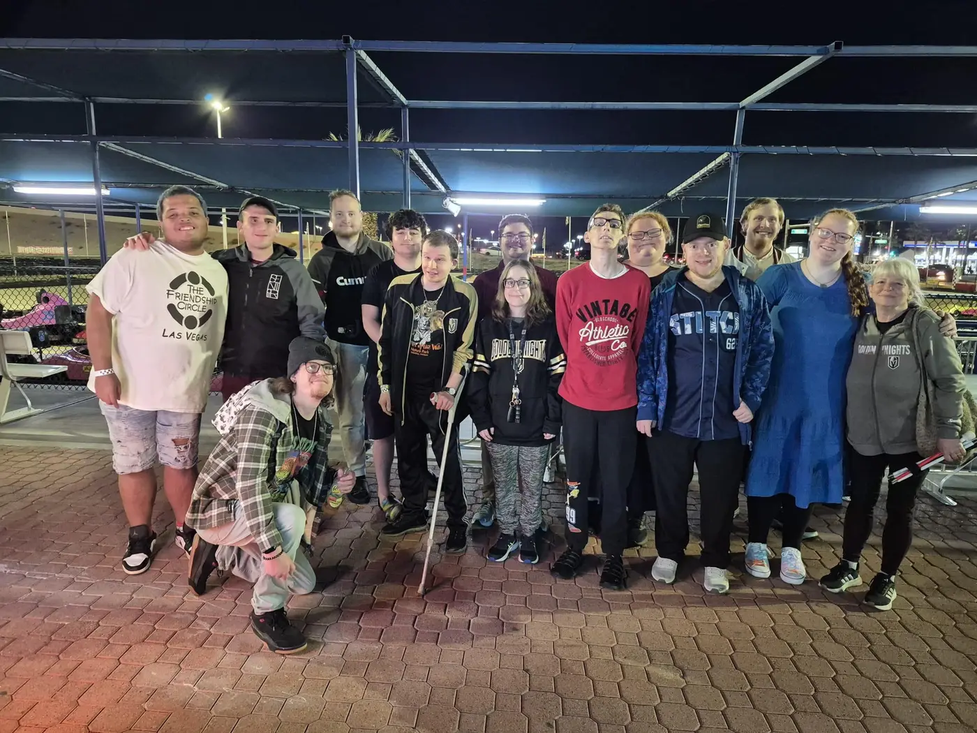 A diverse group of fifteen smiling people pose for a photo at night on a paved surface.