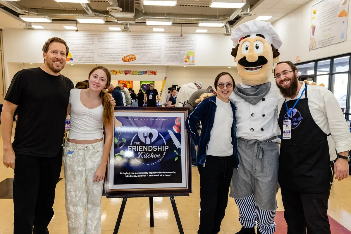 A man, two women, a chef mascot, and a man in an apron pose next to a sign for "The Friendship Kitchen."