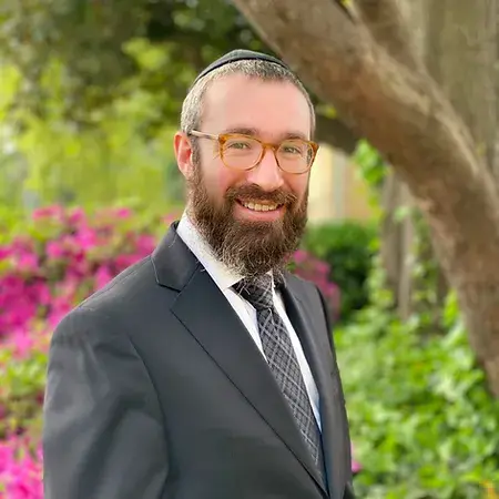 Smiling man in a suit standing outdoors with flowers and trees in the background.