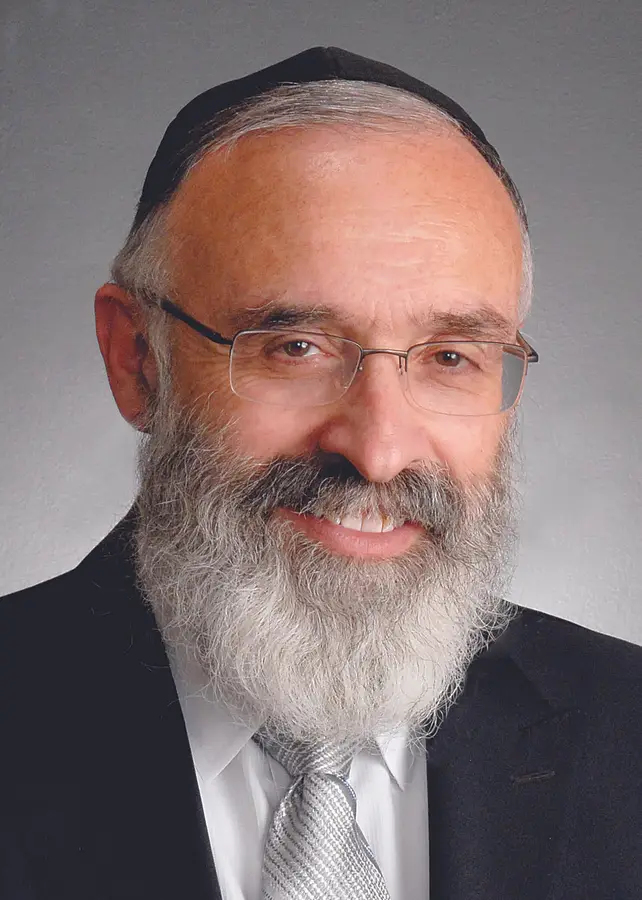 Portrait of a smiling elderly man with a white beard, glasses, wearing a black hat, black suit, white shirt, and a light-colored tie.
