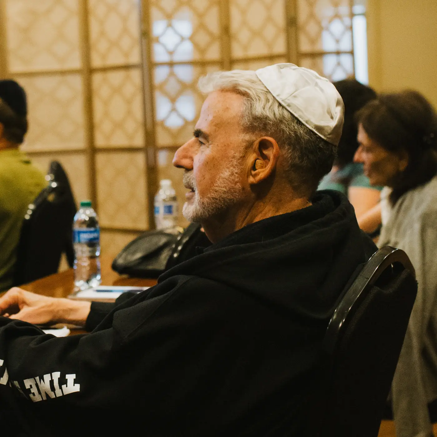 Man with gray hair and beard wearing white kippah, seated at a table during a gathering or conference.