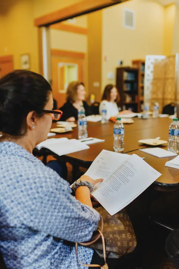 A woman in glasses reading a document at a conference table with other women, bottles of water, and notebooks in a room with warm lighting.