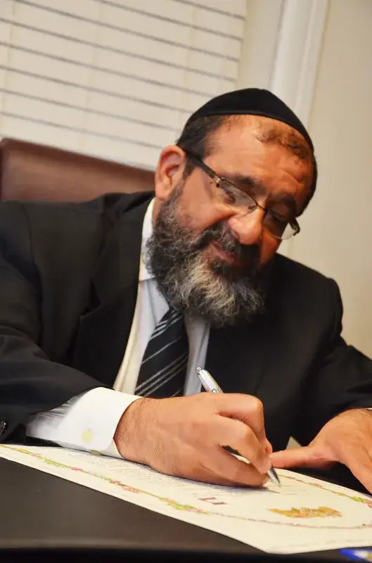 A man in a black suit and striped tie, wearing glasses and a black kippah, sits at a desk, writing or signing documents with a silver pen.