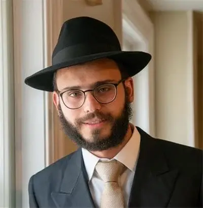 Man with beard and glasses wearing a black fedora hat and a suit with a beige tie, standing indoors near a window.