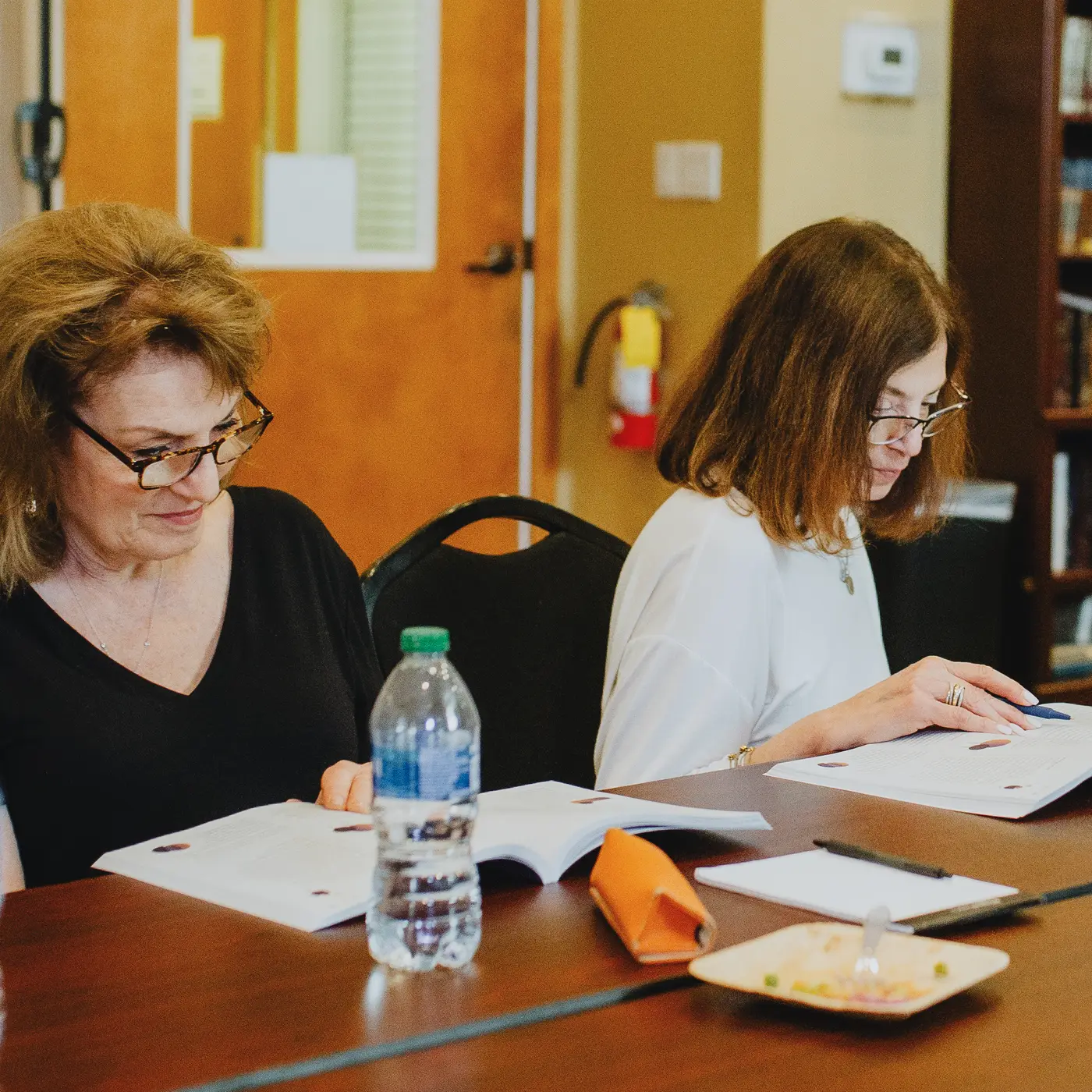 Two women sitting at a table reading documents, with a bottle of water and a bowl of salad in front of them.