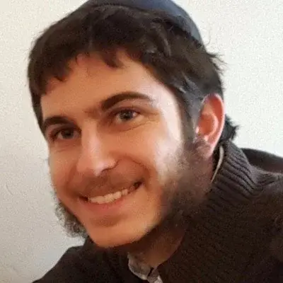 Close-up of a smiling young man with dark hair and beard, wearing a dark sweater, indoors.