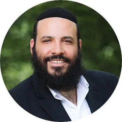Close-up portrait of a man with a beard and mustache, wearing a black hat and suit, smiling outdoors with a blurred green background.