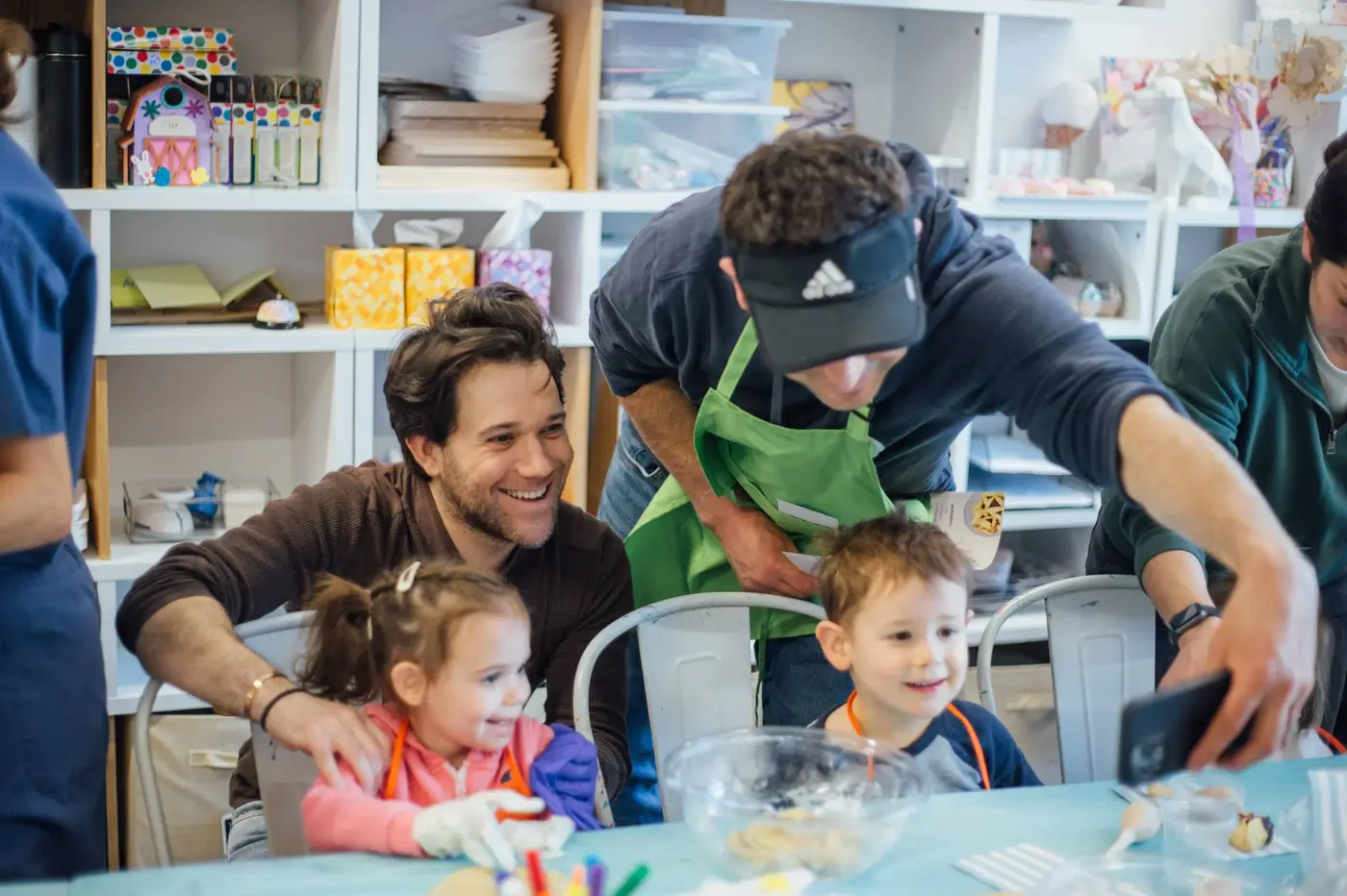 People gathered around a table with children, taking photos, at a festive gathering in a room with shelves filled with craft supplies.