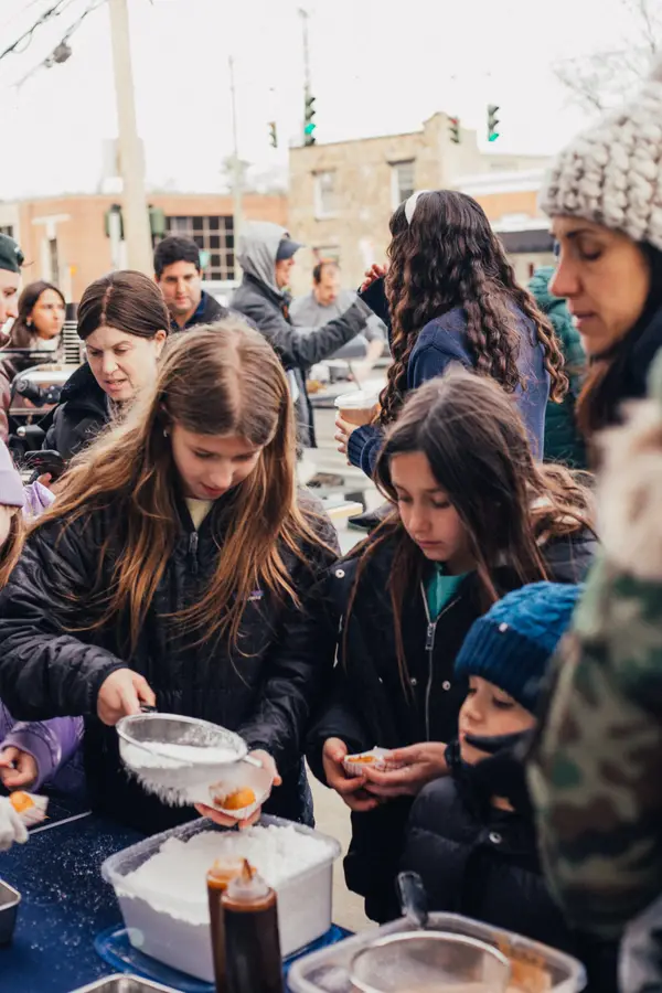 People gathered outdoors around a food stall, serving and receiving food, with a cloudy sky and buildings in the background.