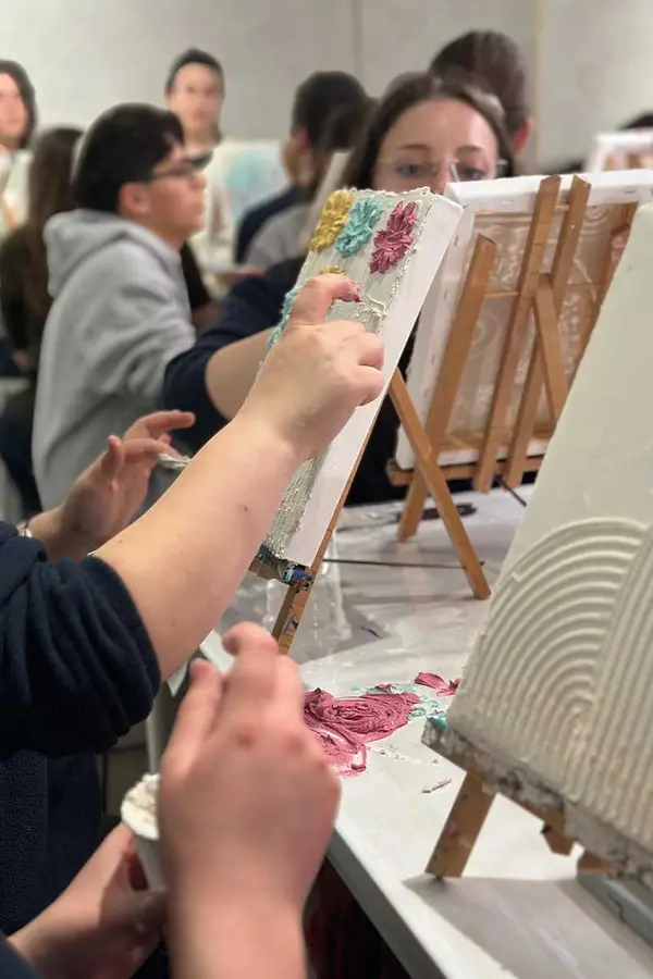 Close-up of hands spreading textured paint on canvases during a group art session, with people painting together in the background.