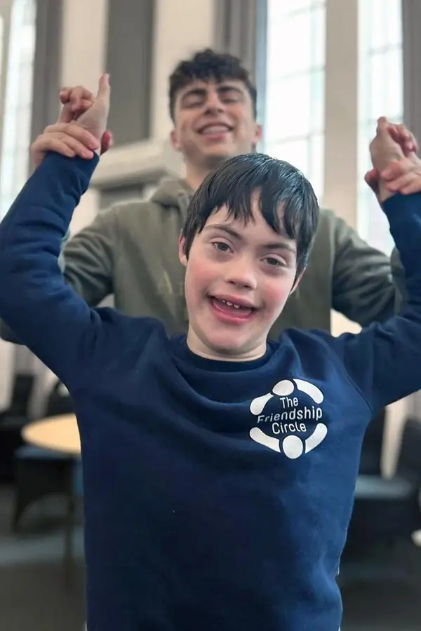 Two males smiling while holding hands up, the one in front wearing a dark blue sweatshirt with The Friendship Circle logo.