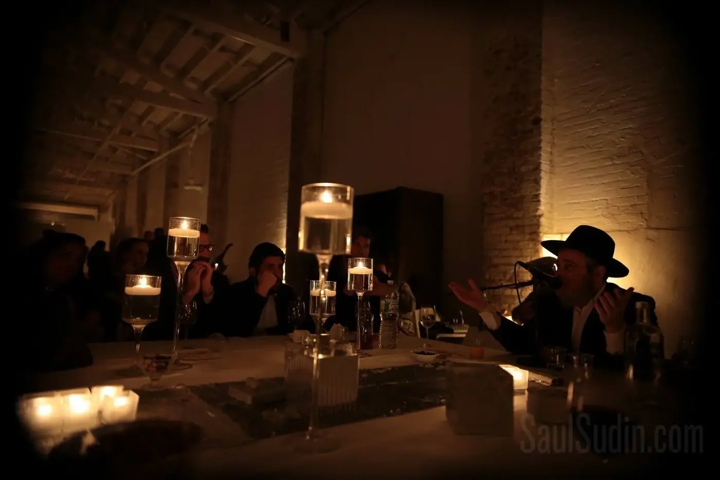 Man wearing a black hat and glasses, speaking into a microphone, gesturing with hands at a dimly lit dinner gathering.