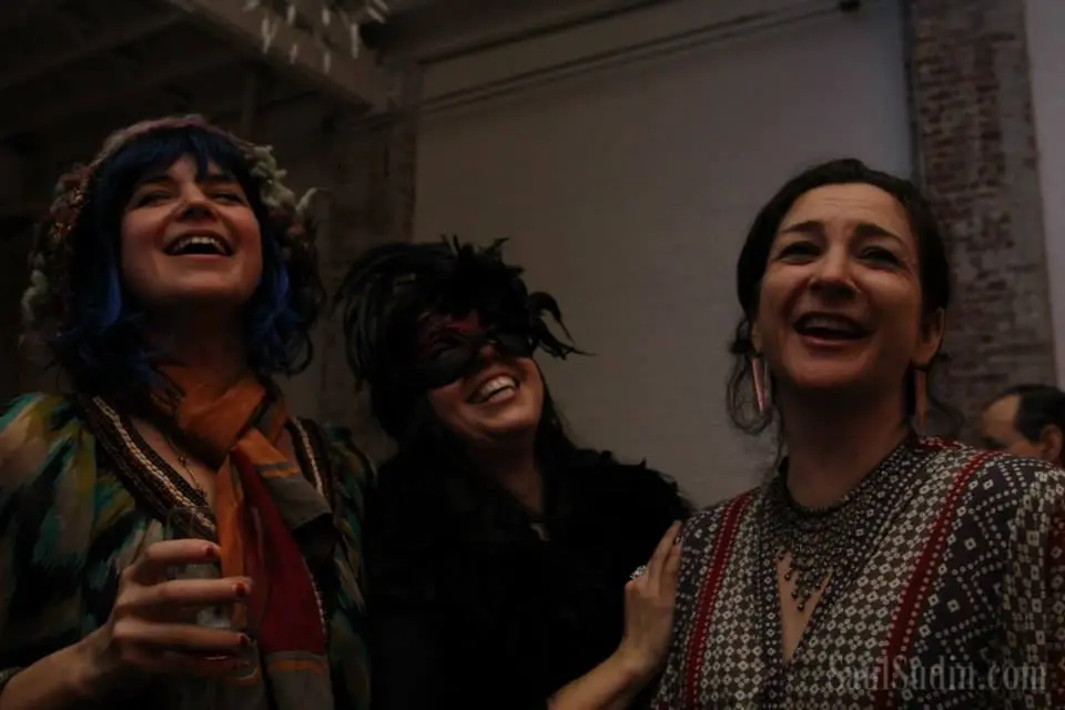 Three women smiling and enjoying themselves at a social event, one holding a glass of beverage, in a dimly lit indoor setting.