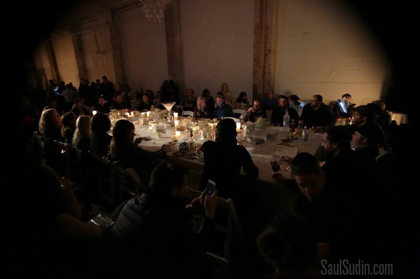 A large group of people sitting around a rectangular dining table with candles, flowers, and drinks in a dimly lit room.