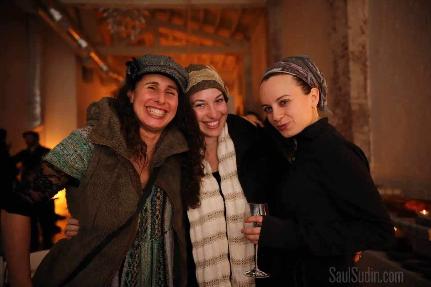 Three women smiling and posing together at an indoor event, one holding a glass of champagne, in a warmly lit rustic setting.
