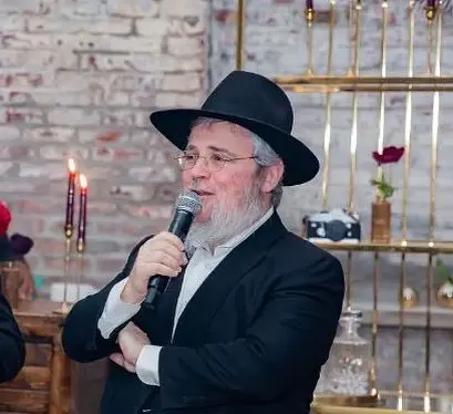 A man with gray hair and beard, wearing glasses, a black hat, and a black suit, speaking into a microphone at a social event.