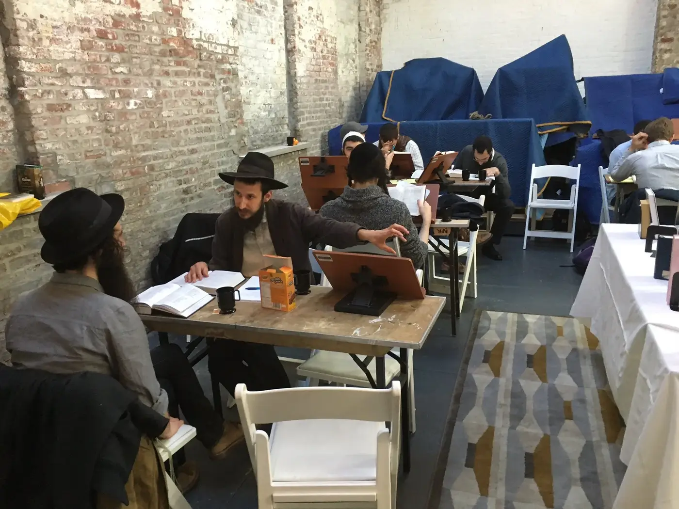 A group of people studying and working in a cafe or study space with brick walls and blue covered items in the background.