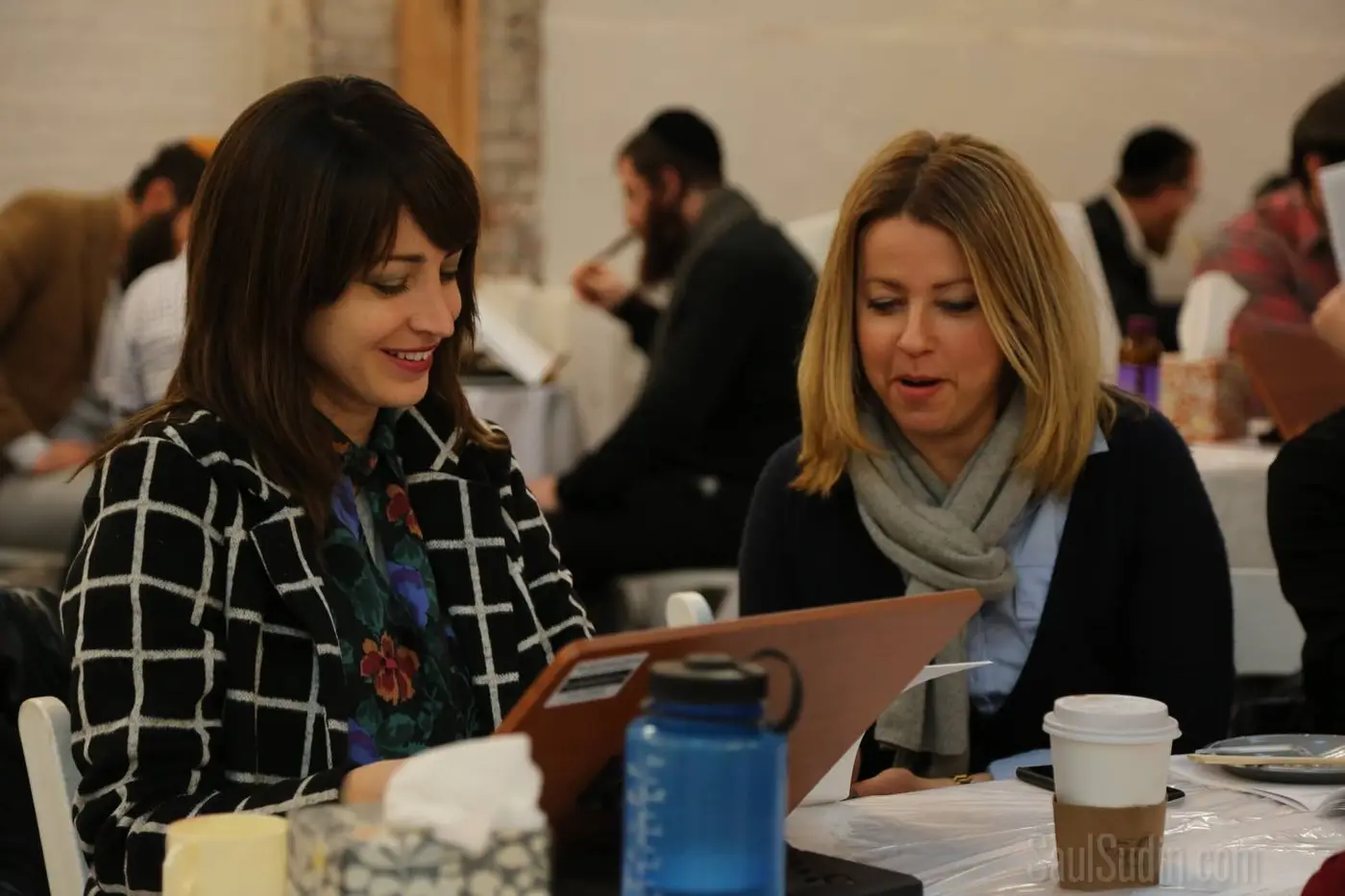 Two women sitting at a table, looking at a menu or document, engaging in conversation in a busy restaurant or cafe.